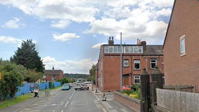 A residential road, flanked by redbrick terraced houses on one side and trees on the other.