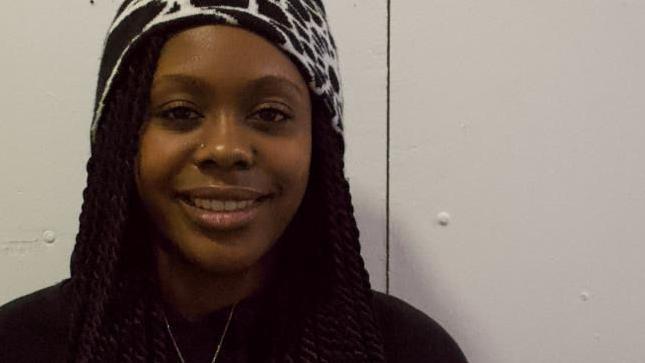 Carrisa Hawkins smiles at the camera, she is wearing a dark coloured top with a gold chain. She has a nose piercing and wearing a brown and white headband in her hair. 