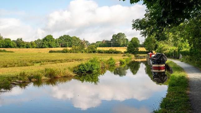 A red, black and cream canal boat on still water with the clouds reflected in it and grassy areas on either side of the water, with a few trees scattered about