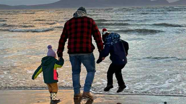 Llun o draeth gyda machlud haul. Mae'r dyn sy'n sefyll ar y tywod yn wynebu i ffwrdd o'r camera, ac yn dal dwylo plant naill ochr iddo.