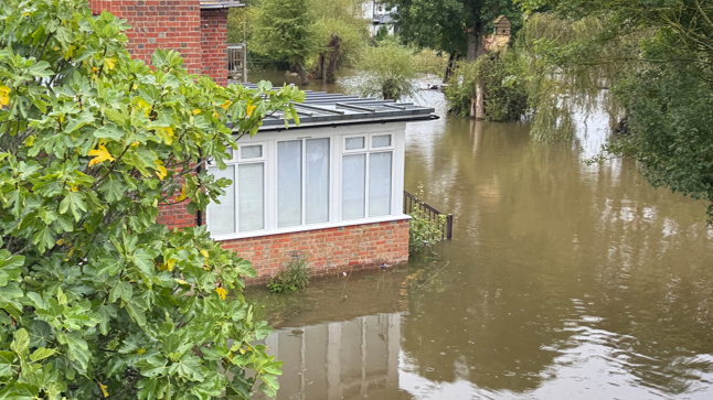 Murky brown floodwater has risen to just below a house's windows. The house has orange bricks and there is a tree to its left. The property is completely surrounded by a lake of water.