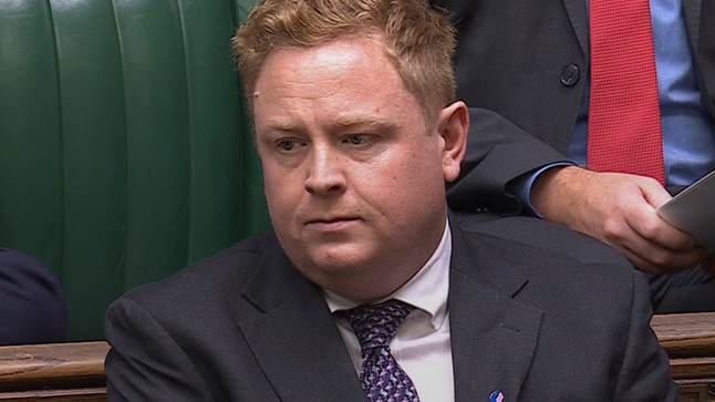A man with red hair and wearing a dark grey suit sits in the House of Commons. He is pictured sat on a green leather seat.