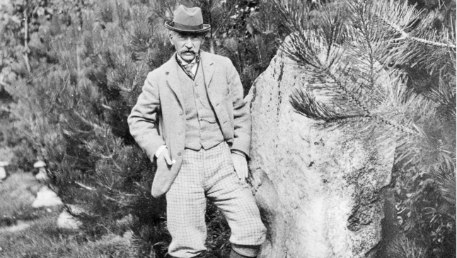 Black and white photo of Thomas Hardy posing next to one of the large, wide sarsen stones which is nearly as tall as him. He has a large moustache and is wearing a trilby hat, a tie, waistcoat and jacket and his hands are in his pockets.