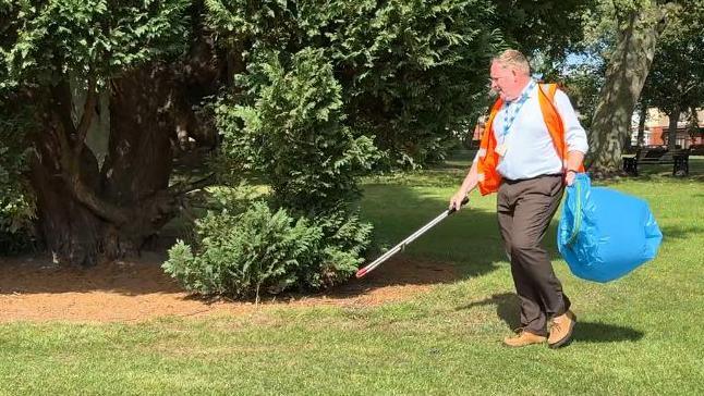 A man wearing work trousers and a shirt, with a high-visibility jacket, carrying a litter picker and a large blue bin bag, collecting rubbish in a park in Shildon.