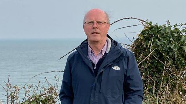 A grey haired man wearing square-shaped glasses and a navy North Face coat looks at the camera while standing in front of some shrubbery and water