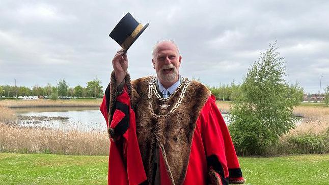 Tom Wootton with short white hair and beard, wearing red and brown mayoral robes and a gold chain. He is holding aloft a black top hat with a gold ribbon around it. There is a nature reserve behind him with reeds, trees and hedges around it.