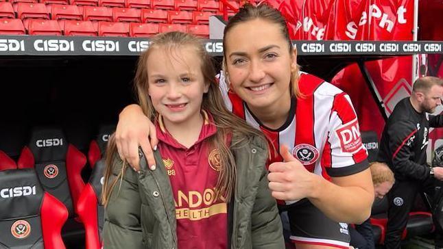 Nevaeh and Maddy on the day she was a Sheffield United mascot. Maddy, wearing her red and white striped kit, has her arm around Neveah's shoulder and gives a thumbs up to the camera. They are both smiling, Neveah missing some of her front teeth. 