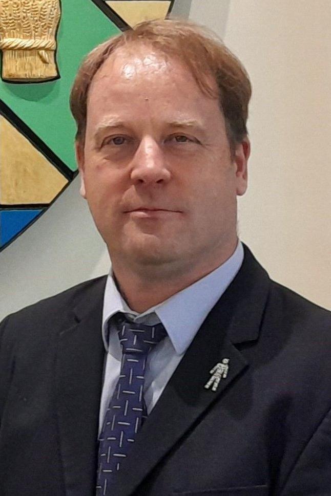A man with short mousey brown hair in a black blazer and a blue tie with white lines on it and a blue shirt. He is smiling into the camera in the head-and-shoulders shot.