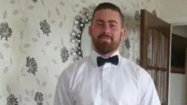 Man in white shirt and bow tie with mirror and decorated wallpaper and brown door behind.