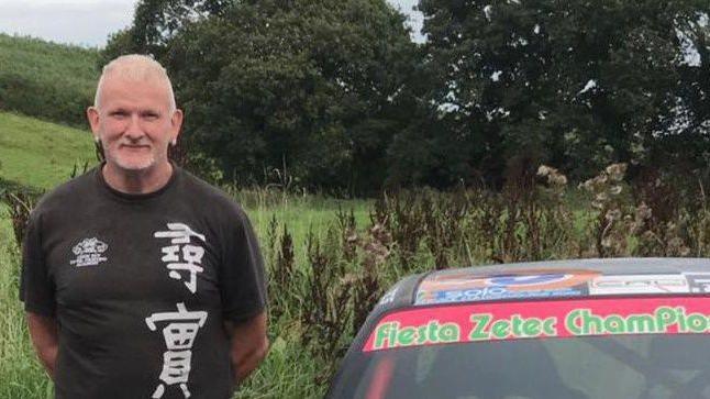 Malcolm McKeown, has short, thinning grey hair and a goatee beard and ear-rings in each ear. He is wearing a black t-shirt with Chinese or Japanese symbols on it and is standing beside a sports car 