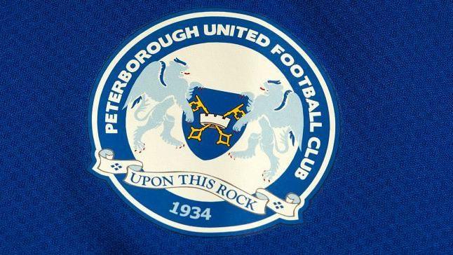 Peterborough United Football Club crest, featuring lions, cross keys, "UPON THIS ROCK" motto and 1934, on a blue background.