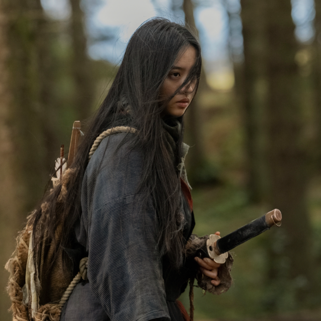 A woman with long black hair and wearing a dark shirt holds a samurai sword