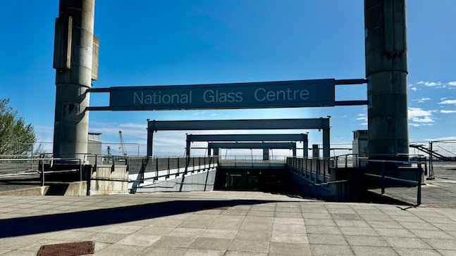 National Glass Centre in Sunderland. Two concrete pillars stand on either side of the centre's wide entrance, which runs into the ground. A sign with the centre's name hangs between the two pillars.