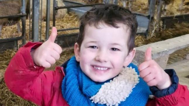 Maldwyn Gwern Evans smiles and sticks both thumbs up to the camera. He is wearing a red jacket and blue scarf