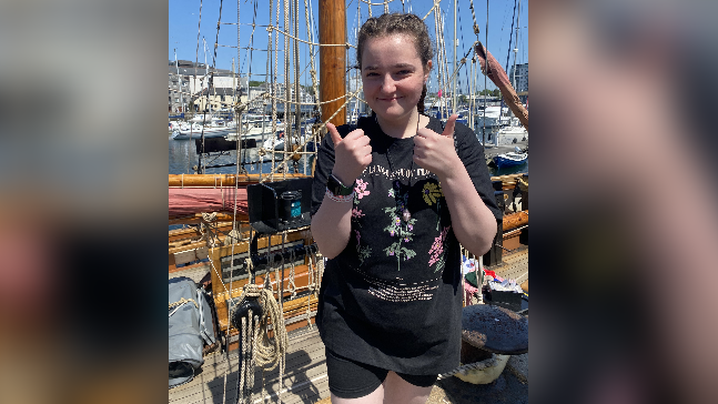 Libby before she had her seizure. She has her thumbs up at the camera and is standing on a boat smiling at the camera on a sunny day in a dock.