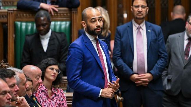 A man in a cobalt coloured suit and a red tie standing up and talking in parliament.