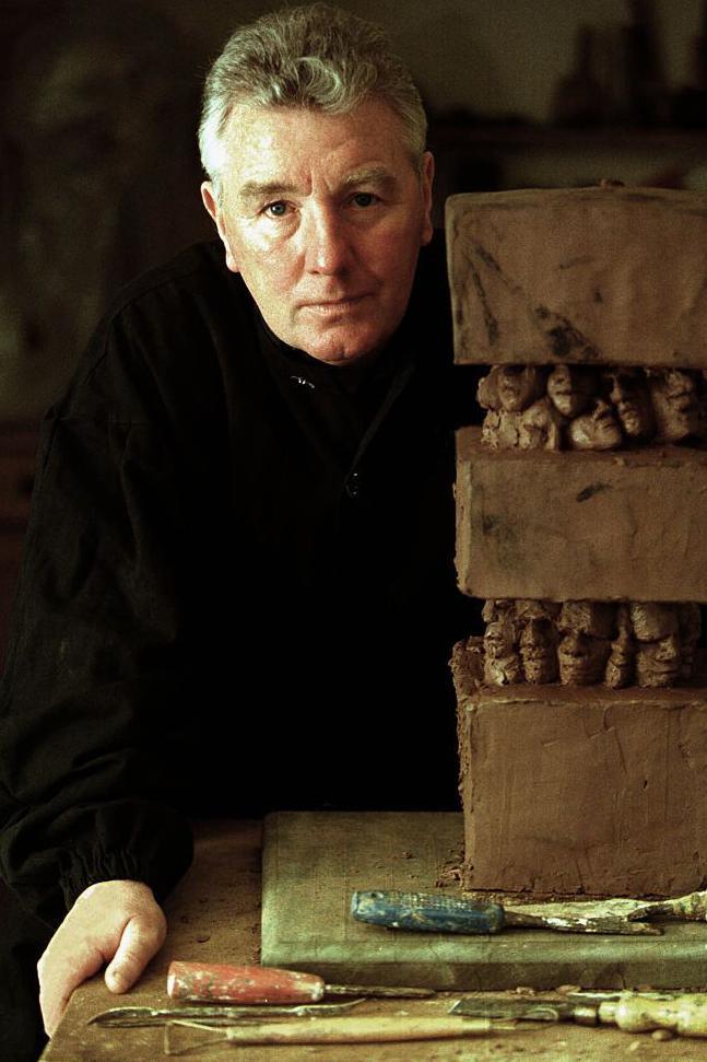 Jimmy Boyle, wearing black clothing, standing next to a three-level clay sculpture. It is placed on a table, with tools lying nearby.