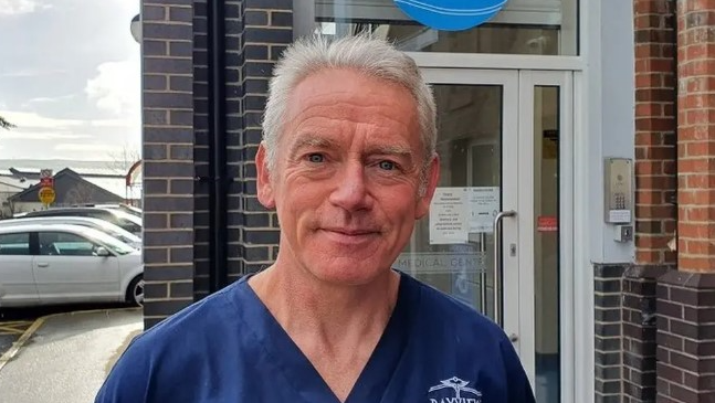 Shows a man with grey hair in a blue v-neck top standing in front of a building and cars to the left
