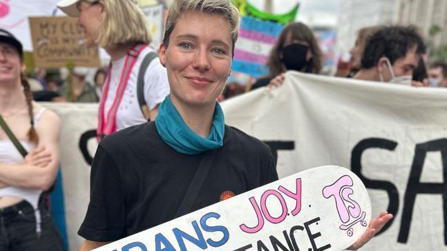 An attendee of the event holding a sign, of which you can just see the words "trans" written in blue and "joy" written in pink against a white background