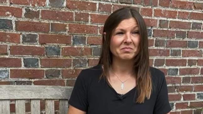 A woman wearing a black shirt. She is sat on a bench in front of a brown brick wall.