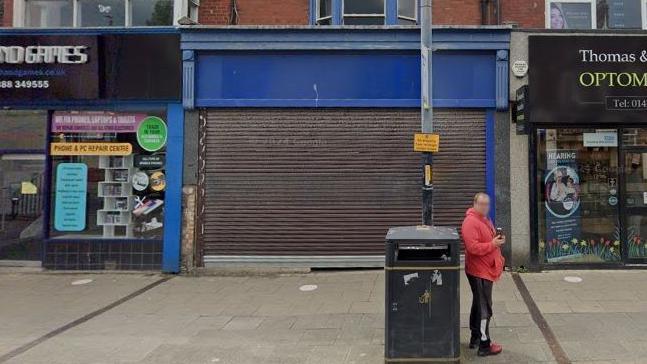 A Google Street View screenshot of the Screams Court 2.0 venue in Hartlepool. The unit has its shutter down and has a blue panel without a sign. It is flanked by shops. A man in a red hoodie and tracksuit bottoms is drinking a can outside.