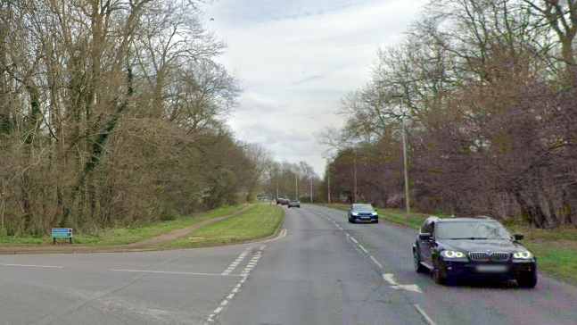 Google maps view of Bretton Way with cars driving on it and a side turning into Barnstock with a blue sign and trees on either side of the road.