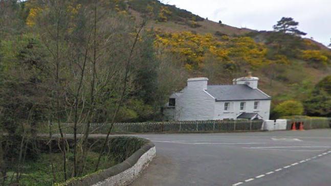 The junction at Ballig Bridge, which has a low wall surrounding trees on the left and a white cottage on the right.