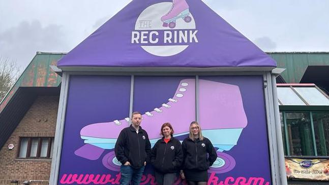 Three people in black coats standing outside a purple building which says "The Rec Rink" and and features a large picture of a purple roller skate.