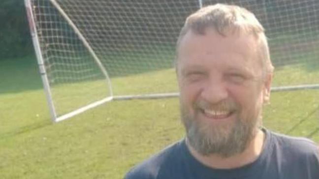 Robin Reed smiling for the camera. He has a grey beard and is wearing a navy blue t-shirt that has the Labretta brand. Behind him is a football goalpost.