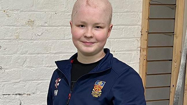 Finn wearing a navy blue cricket tracksuit standing in front of a brick wall painted white. He is smiling.