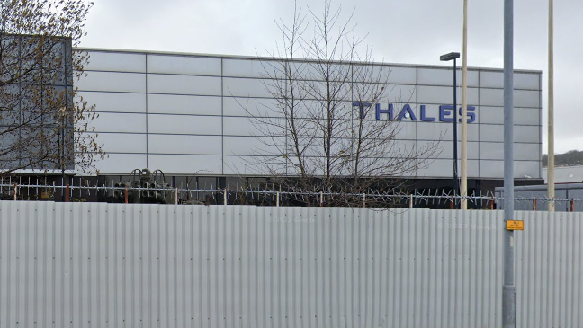 THALES title is top right in dark blue. The building is made up of reflective windows and has high walled security. 