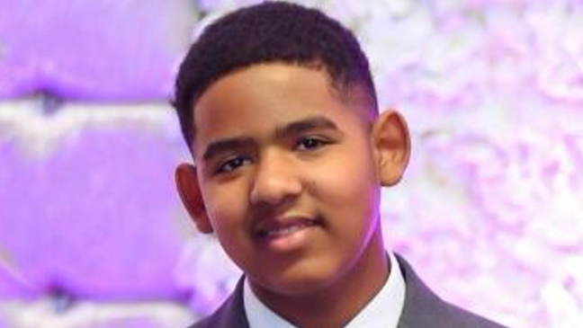 Yassin is smiling at the camera and in a grey suit and white shirt and has short, dark hair. There is a purple patterned background behind him.