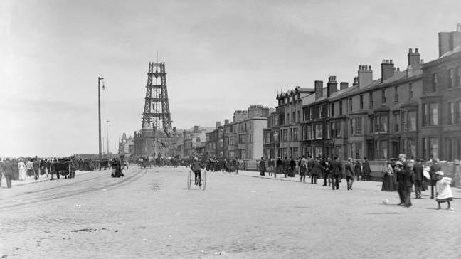 Black-and-white photo of Blackpool Promenade pictured in June 1893 with visitors in the distance near two arc lamps on 60ft poles and Blackpool Tower which is partly built in the distance.