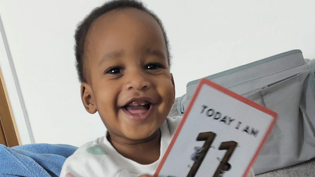 A young child with dark hair wearing a white T-shirt with coloured spots on it is smiling. He is holding a card which reads: "TODAY I AM 11," although the bottom of the card is cut off.