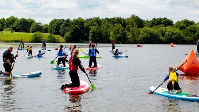 A colourful array of paddle-boards and paddle-board students wearing life jackets, seen from the back, the lake stretching out in front of them.  The background is the grassland and trees that surround the lake.