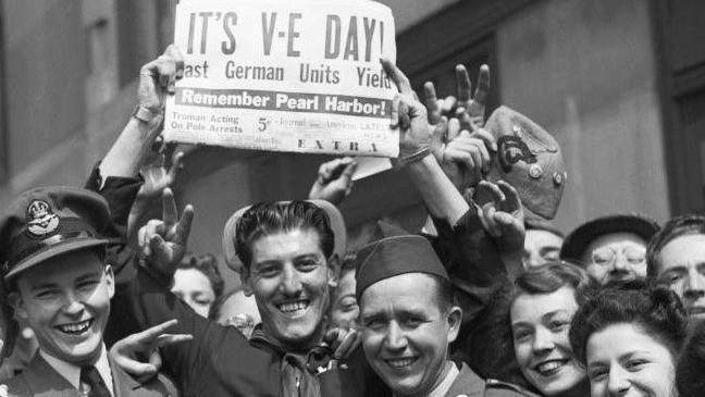 Man in a crowd of smiling faces holds up a newspaper