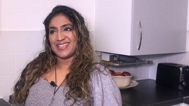 Sunita Kaur Singh sits in the kitchen of her new social home with a cupboard behind her and a toaster and fruit bowl on the worktop. She has long brown hair past her shoulders and is smiling.