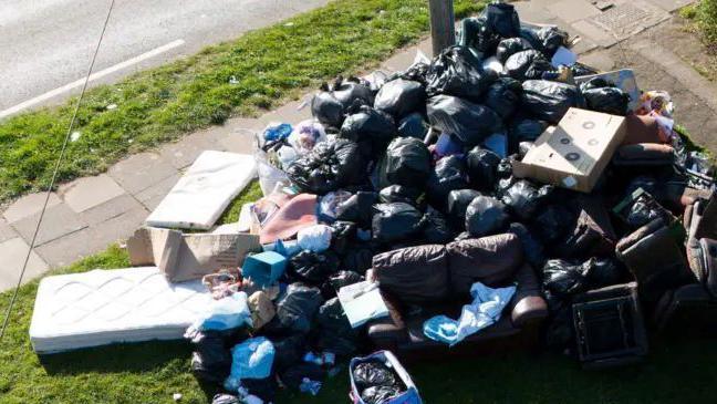 A large pile of black bin bags, with some blue bags and mattresses, cardboard and other waste.