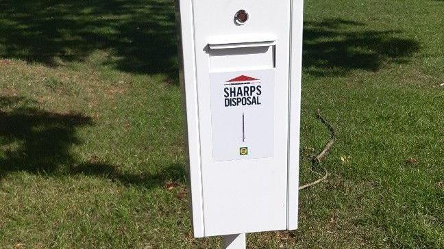 A white needle bin- that looks like a small letter box- placed on the grass.