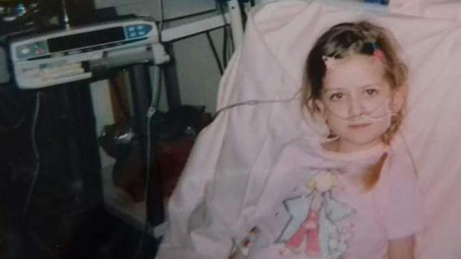A young girl sits in a hospital bed, wearing a pink nightie with a cartoon fairy design on it. She has tubes attached to her face. She has brown hair and is smiling slightly. Hospital equipment can be seen in the background.