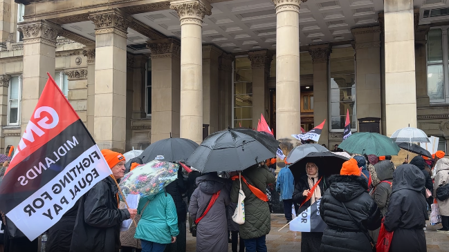 Dozens of people standing outside a large building. They are holding umbrellas and GMB union flags