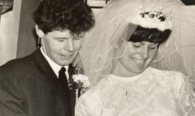 Donald and Margaret Tyman on their wedding day. Donald has short, black curly hair wearing a black wedding suit while Margaret has short black hair wearing a tiara and veil and white wedding dress and gold cross necklace. Both are smiling.