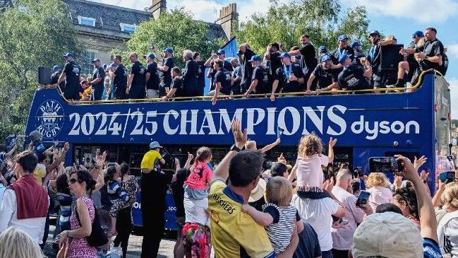 Blue bus with 2024/2025 champions written on the side. Bath players are on the top of the bus dressed in black t shirts with blue Bath caps. Lots of people are waving and taking photos in the foreground.