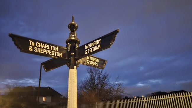 Black finger signage pointing in four different directions. The signs are on a white post. The sky is dark grey behind the signs