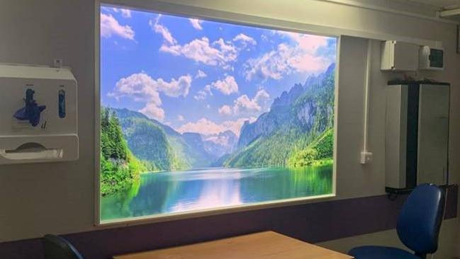 Screen with a shot of a lake which is surrounded by mountains and trees. Light is reflecting on the lake with fluffy clouds visible in the shot.