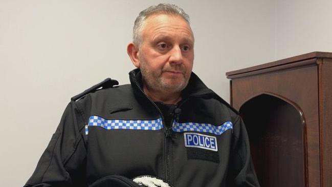 Damian Kitchen pictured wearing a black police uniform. He is also holding his police hat in his hands and standing next to a wooden cabinet.