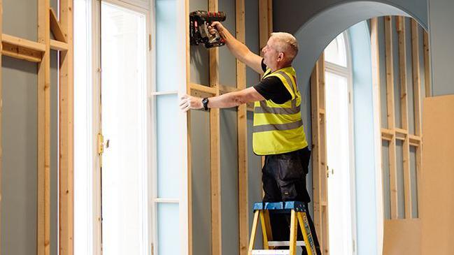 A man in a high visibility jacket stands on top of stepladders nailing wood battens onto a wall with a nail gun. Around him are other wood panels and power tools.