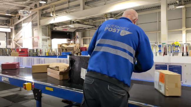 A man wearing a Guernsey Post jacket putting parcels on the moving conveyor belt in the warehouse.