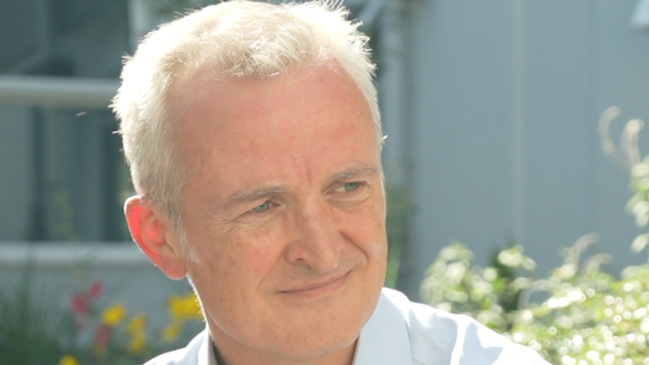 Dr Ewen Cameron smiles while looking away from the camera. He sits outside where bushes and flowers can be seen behind him. He has short grey hair and wears a white shirt. 