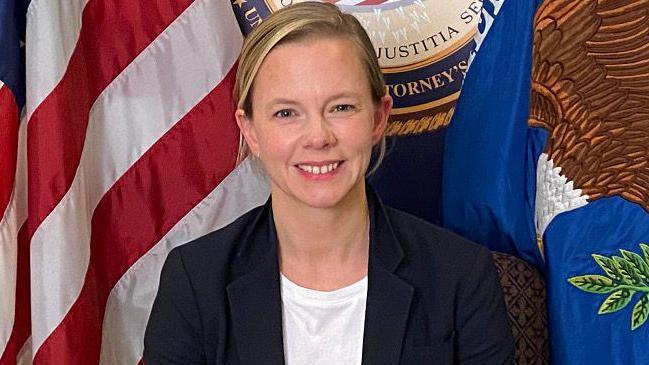 A woman with short blonde hair looks into the camera with a broad smile. She is wearing a fark jacket and white t-shirt. In the background we can partially see the flag of the United States, the flag of the US Department of Justice and the seal of the US Department of Justice. 